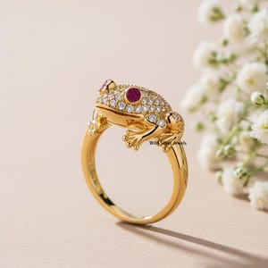 Puede incluir: Un anillo de rana dorado con ojos de rubí y un cuerpo incrustado con pequeñas piedras preciosas transparentes. El anillo presenta un diseño detallado de rana con piel texturizada y patas delicadas. El anillo está sobre un fondo suave y neutro.