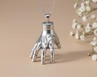 May include: A silver-colored pendant necklace featuring a stylized hand with visible stitching details. The hand is suspended from a delicate chain. The text "Wild Silver Jewels" is visible on the hand.