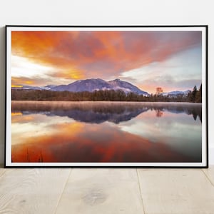 May include: A scenic photograph of a mountain lake with a reflection of the sky and clouds in the water. The sky is a vibrant orange and pink, with a few wispy clouds. The mountain is covered in trees and there is a small forest in the foreground.