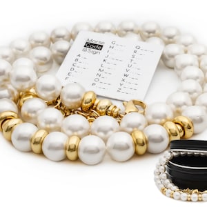 Könnte beinhalten: Eine doppelreihige Perlenkette mit goldenen Akzenten und einem Verschluss. Die Kette befindet sich in einer kleinen Blechdose auf weißem Hintergrund. Die Dose enthält auch eine kleine Karte mit dem Morsealphabet.