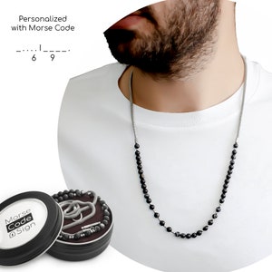 May include: A black beaded necklace with a silver chain, worn by a person. The necklace is in a round tin box with the words "Morse Code Sign" on the lid.