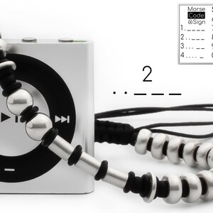 May include: A silver and black beaded necklace with a silver iPod Nano. The necklace features a silver bead with a black rubber band around it. The iPod Nano has a black circle in the centre and a play button on the left and a fast forward button on the right. The Morse code for the number 2 is shown below the iPod Nano.