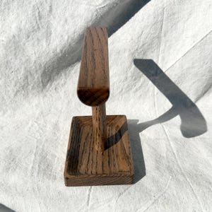 Puede incluir: Un expositor de joyas de madera con base rectangular y parte superior vertical curvada. El expositor es de madera marr&oacute;n claro con veta visible. Dise&ntilde;ado para sujetar pulseras o collares.