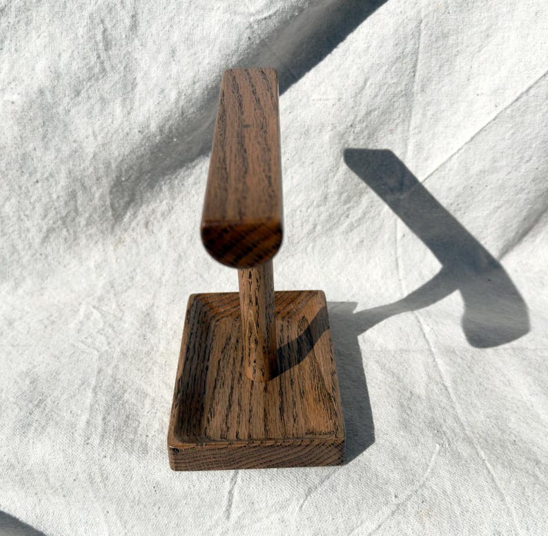 Puede incluir: Un expositor de joyas de madera con base rectangular y parte superior vertical curvada. El expositor es de madera marr&oacute;n claro con veta visible. Dise&ntilde;ado para sujetar pulseras o collares.