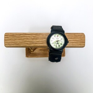 Puede incluir: Un reloj Swiss Army negro con esfera blanca y detalles verdes, con una correa de tela negra. El reloj se exhibe en una barra de madera marr&oacute;n claro con un peque&ntilde;o gancho de lat&oacute;n.