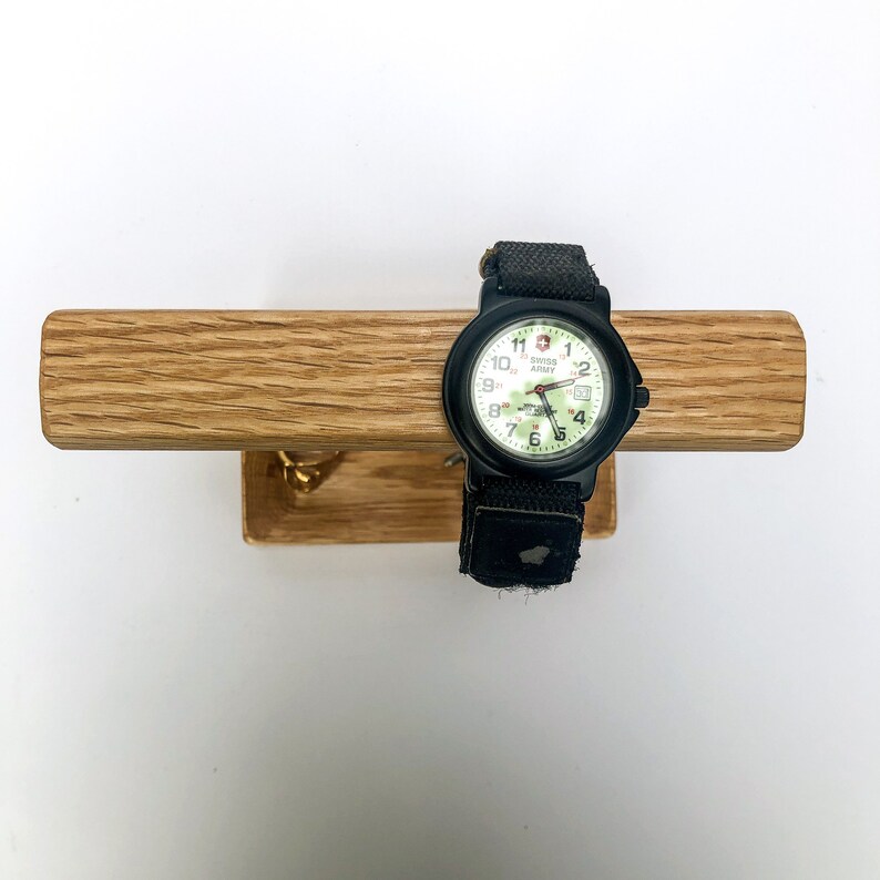Puede incluir: Un reloj Swiss Army negro con esfera blanca y detalles verdes, con una correa de tela negra. El reloj se exhibe en una barra de madera marr&oacute;n claro con un peque&ntilde;o gancho de lat&oacute;n.