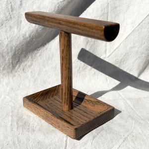 Puede incluir: Un expositor de joyas de madera con una base rectangular y un poste vertical que soporta una barra horizontal. El expositor es de madera marr&oacute;n oscuro con veta visible, adecuado para exhibir collares o pulseras.