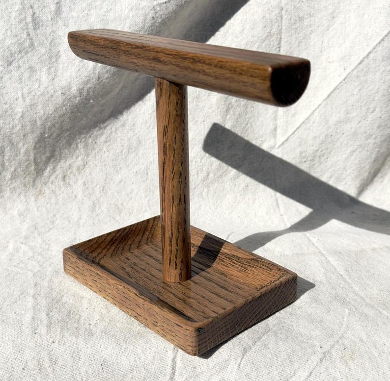 Puede incluir: Un expositor de joyas de madera con una base rectangular y un poste vertical que soporta una barra horizontal. El expositor es de madera marr&oacute;n oscuro con veta visible, adecuado para exhibir collares o pulseras.