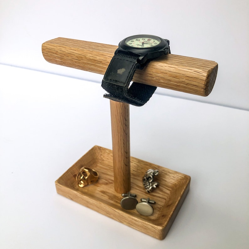 Puede incluir: Un expositor de madera para relojes y joyas. El soporte tiene una barra horizontal para un reloj, un soporte vertical y una base de bandeja. Se exhiben un reloj negro, un anillo de calavera dorado y gemelos plateados.
