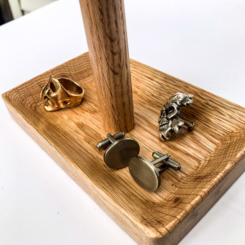 Puede incluir: Una bandeja de madera con un poste central de madera sostiene un anillo de calavera dorado, un gemelo de calavera plateado y dos gemelos ovalados de bronce. La bandeja tiene un acabado de vetas de madera natural.
