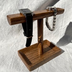 Puede incluir: Un soporte de reloj de madera con una base rectangular y una barra horizontal. Un reloj negro con caja plateada y un reloj plateado con correa plateada se exhiben en el soporte. El soporte est&aacute; hecho de madera de tono medio.