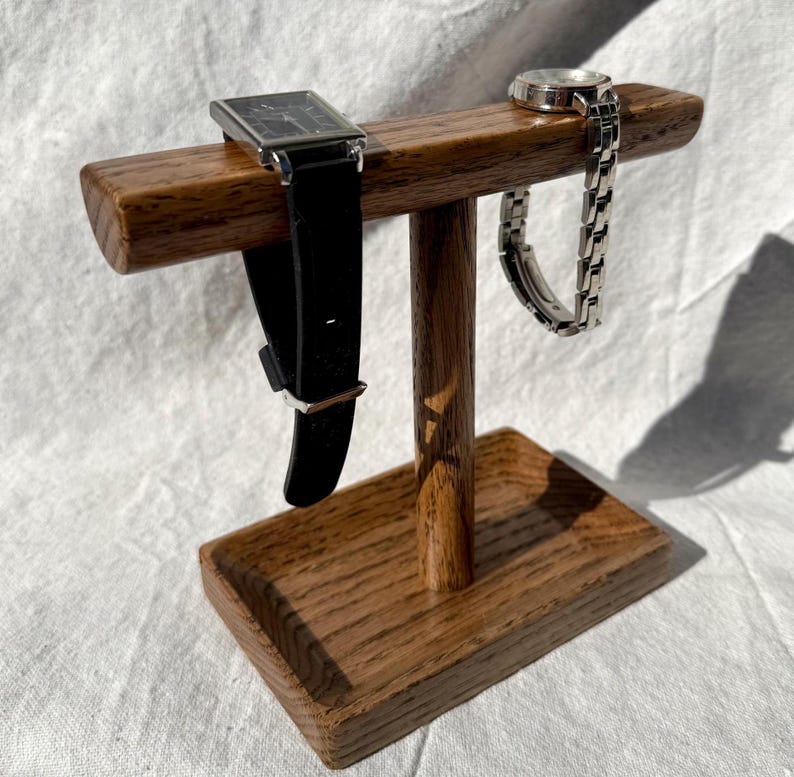 Puede incluir: Un soporte de reloj de madera con una base rectangular y una barra horizontal. Un reloj negro con caja plateada y un reloj plateado con correa plateada se exhiben en el soporte. El soporte est&aacute; hecho de madera de tono medio.