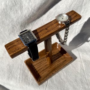 Puede incluir: Un soporte de madera para relojes que muestra dos relojes. Uno tiene una esfera rectangular negra y una correa de cuero negro. El otro tiene una correa plateada y una esfera redonda. El soporte es de madera marr&oacute;n claro.