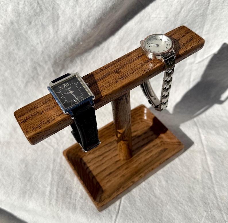 Puede incluir: Un soporte de madera para relojes que muestra dos relojes. Uno tiene una esfera rectangular negra y una correa de cuero negro. El otro tiene una correa plateada y una esfera redonda. El soporte es de madera marr&oacute;n claro.