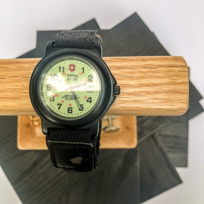 Puede incluir: Reloj negro Swiss Army con esfera verde claro y correa de lona negra. El reloj tiene n&uacute;meros y manecillas blancas, con el logotipo de Swiss Army. Se muestra sobre un soporte de madera.