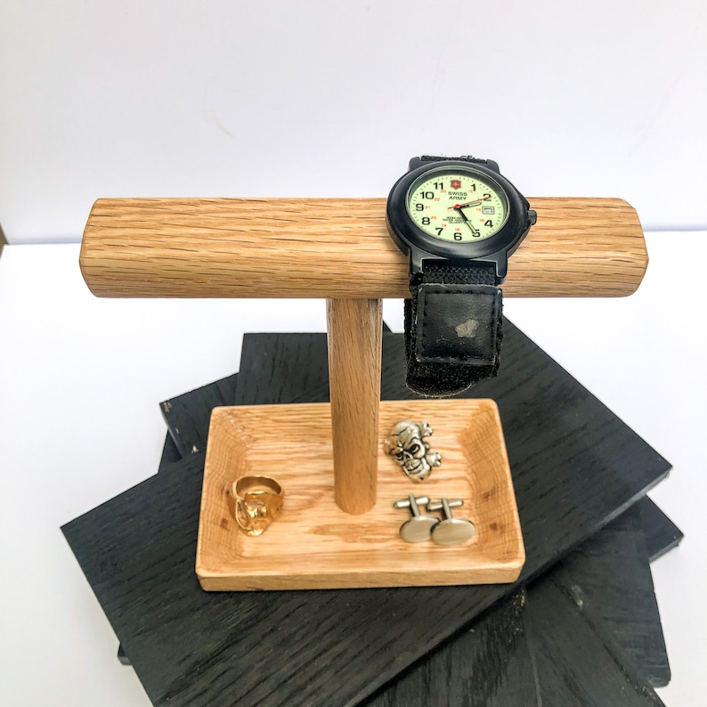 Puede incluir: Un expositor de madera para relojes y joyas. El expositor tiene una barra horizontal para un reloj, una peque&ntilde;a bandeja para anillos y gemelos, y un reloj negro con una correa negra. Un anillo dorado, gemelos de calavera y un anillo de calavera est&aacute;n en la bandeja.