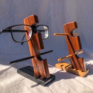 Puede incluir: Dos soportes de madera para gafas. Un soporte tiene un par de gafas con montura negra. Los soportes son de madera marrón oscuro con una base negra. El otro soporte está vacío. Los soportes están diseñados para sujetar gafas.