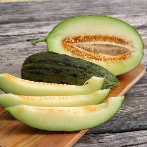 May include: A green melon cut in half, revealing the light yellow flesh and seeds. Four slices of melon are arranged on a wooden chopping board.