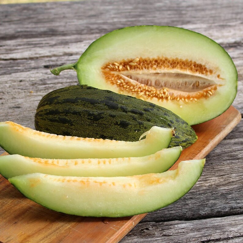 May include: A green melon cut in half, revealing the light yellow flesh and seeds. Four slices of melon are arranged on a wooden chopping board.