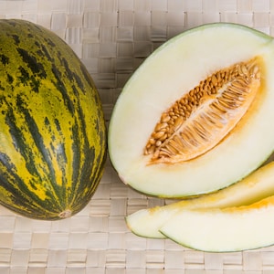 May include: A whole green and yellow striped cantaloupe melon next to a half cantaloupe melon with seeds and two slices of cantaloupe melon.