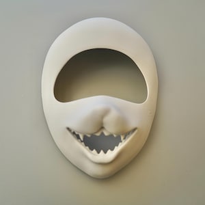 May include: A white ceramic mask with a large, open mouth and sharp teeth. The mask has a hole for the eyes and a small nose.