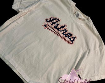 May include: White t-shirt featuring the word "Astros" in a stylized script with a star and a red and navy blue color scheme. The shirt is a short-sleeved crew neck.