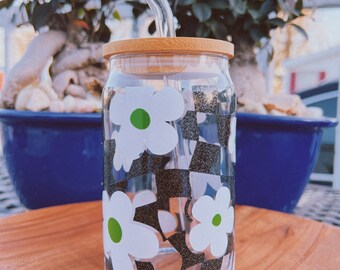 May include: A clear glass can-shaped tumbler with a bamboo lid and a curved glass straw. The tumbler features a black and white checkered pattern with white daisy flower graphics with green centres. The tumbler sits on a wooden surface.