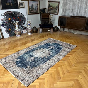 May include: Rectangular, vintage-style rug with a central blue and white design, surrounded by a border of beige and brown. The rug is placed on a wooden floor with a herringbone pattern, and is in a room with paintings and decorative vases.