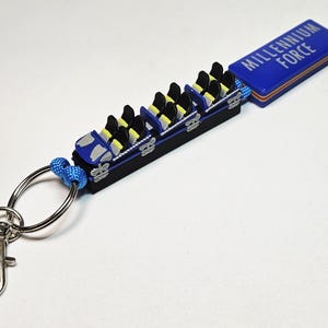 Puede incluir: Un llavero de montaña rusa azul y negro con el texto "MILLENNIUM FORCE" en una etiqueta rectangular azul. El llavero presenta un tren de montaña rusa en miniatura con asientos negros y reposacabezas amarillos, sujeto a un llavero y cierre plateados.