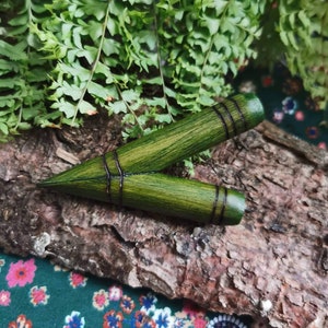 Pode incluir: Uma flecha de madeira verde com listras pretas, apoiada em um pedaço de casca de árvore. A flecha é pontiaguda e tem uma ponta arredondada.