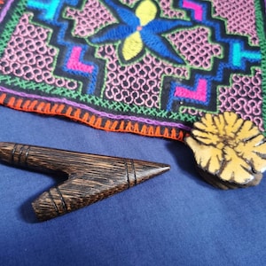 May include: A dark brown wooden Y-shaped object, a yellow and brown floral stamp, and a colourful embroidered textile with geometric patterns are displayed on a blue surface. The textile features pink, blue, and green stitching.