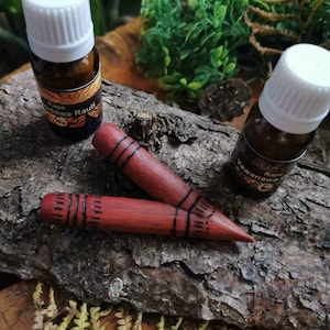 Könnte beinhalten: Zwei kleine braune Glasflaschen mit weißen Kappen und Etiketten mit der Aufschrift "Rape Yawana Rauti" und "Rape Yawanaawa". Zwei Holzstäbchen mit roter Farbe und schwarzen Markierungen liegen auf einer braunen Holzoberfläche.