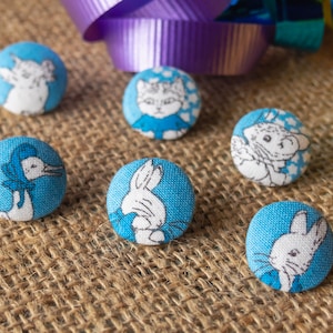 May include: Six blue fabric-covered buttons with white animal illustrations. The buttons feature a cat, bunny, and other animal designs. The buttons are on a hessian surface with a purple ribbon in the background.