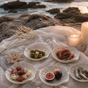 Könnte beinhalten: Fünf weiße Keramikschalen in Form von Jakobsmuscheln, die jeweils verschiedene Lebensmittel wie Oliven, Prosciutto, Caprese-Salat, frische Feigen und Sardinen enthalten, arrangiert auf einem weißen Stofftuch mit zwei brennenden Stumpenkerzen.
