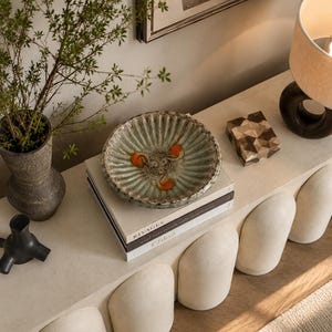 Op de afbeelding: Een decoratieve opstelling met een groene keramische kom met oranje accenten, rustend op een stapel boeken. Een vaas met groen, een geometrische houten doos en een lamp met een beige kap zijn ook aanwezig. De scène is geplaatst op een lichtgekleurd oppervlak.