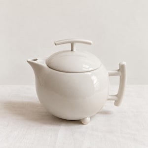 Op de afbeelding: Een glanzende, gebroken witte keramische theepot met een bolvormig lichaam, een korte tuit en een deksel met een T-vormig handvat. De theepot heeft een verticaal handvat aan de zijkant en rust op kleine, afgeronde voetjes met een gladde afwerking.