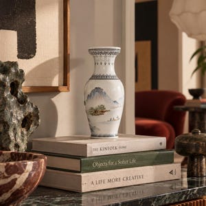Puede incluir: Un jarrón de cerámica blanca con cuello estrecho y borde acampanado, que presenta una pintura de paisaje en azul y verde con montañas y agua. El jarrón se asienta sobre una pila de tres libros titulados The Kinfolk Home, Objects for a Softer Life y A Life More Creative.