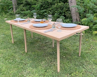 May include: A wooden table with four place settings, each with a white plate, silverware, and a glass. The table is set up outdoors on a grassy lawn with trees in the background.