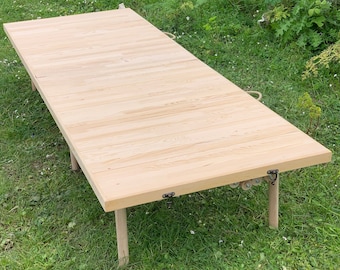 May include: A wooden folding bed frame with a natural finish. The frame is made of slats and has four legs. The frame is folded up and sitting on a grassy surface.