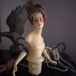 May include: A vintage porcelain doll with painted features and a brown wig. The doll is wearing a pearl necklace and is posed with one arm outstretched. The doll is sitting on a decorative metal base.