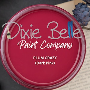 May include: A round container of Dixie Belle Paint Company's Plum Crazy (Dark Pink) paint. The container is a deep magenta color, with the brand name and product name in white text. The image is a top-down view.