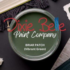 May include: A jar of Dixie Belle Paint Company's Briar Patch (Vibrant Green) paint. The jar is open, revealing the green paint inside. The company logo is in red and white. The jar is surrounded by decorative items.