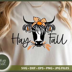 Puede incluir: Una camiseta blanca con una ilustraci&oacute;n en blanco y negro de una vaca con un lazo de lunares. La vaca est&aacute; comiendo tallos de trigo. El texto "Hay Fall" est&aacute; escrito en letras cursivas negras.