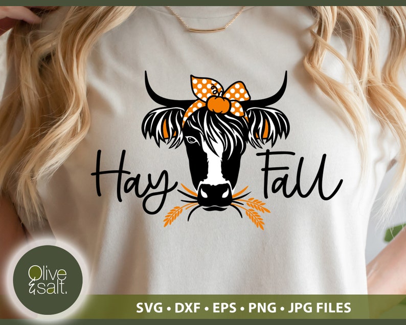 Puede incluir: Una camiseta blanca con una ilustraci&oacute;n en blanco y negro de una vaca con un lazo de lunares. La vaca est&aacute; comiendo tallos de trigo. El texto "Hay Fall" est&aacute; escrito en letras cursivas negras.