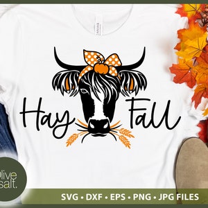 Op de afbeelding: Wit T-shirt met een zwart-witte illustratie van een koe die een stippen strik en een pompoen draagt. De tekst "Hay Fall" staat in zwart onder de koe.