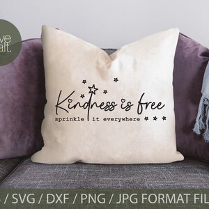 May include: A white throw pillow with the text "Kindness is free sprinkle it everywhere" in black lettering with stars and a heart. The pillow is on a gray couch with a purple throw pillow and a blue throw blanket.