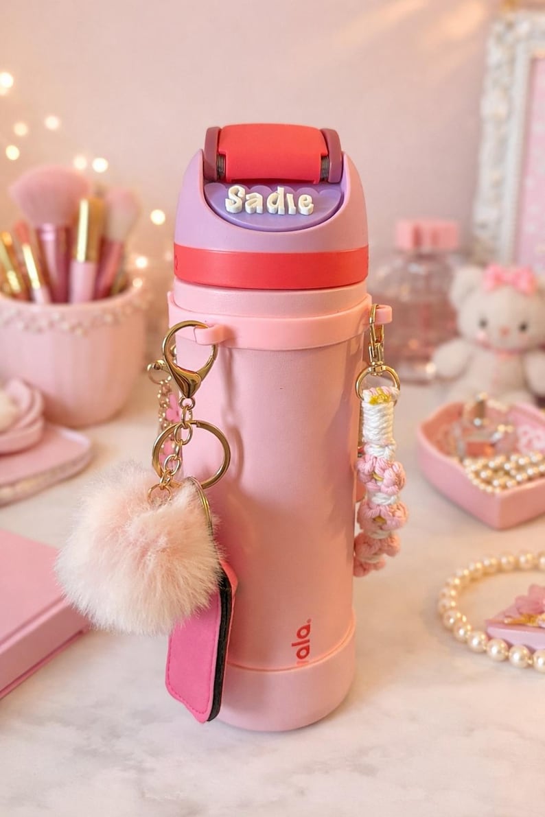 May include: Pink water bottle with a red and purple lid, featuring the name "Sadie". The bottle has a gold keychain with a pink fluffy pom-pom and a pink tag. A decorative pink and white macrame keychain is also attached.