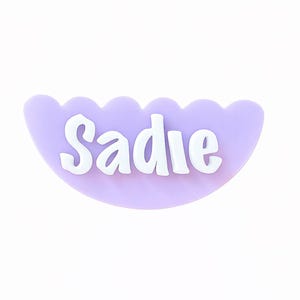 May include: A light purple, scalloped-edge name tag with the name "Sadie" in white, bold letters. The name tag is a half-circle shape, and the background is plain white.