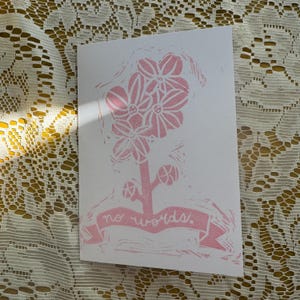 May include: A white greeting card with a pink floral design and the words "no words." The illustration features a stylized flower with multiple blooms and a banner. The card is on a textured, yellow-toned surface.