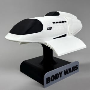 Può includere: Un modello di navicella spaziale bianca e nera con il testo "B229" stampato sul lato. La navicella spaziale è montata su un supporto nero con la scritta "BODY WARS" in lettere nere su sfondo grigio.